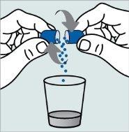 Ręce otwierają niebieską kapsułkę z granulkami spadającymi do szklanki z przezroczystym płynem