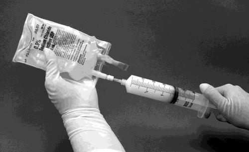 Mano con guanto che collega una siringa piena a una sacca IV trasparente con etichetta e soluzione chiara