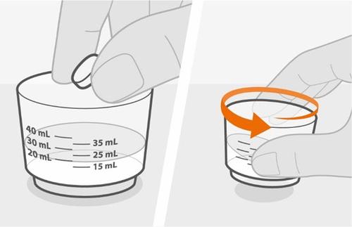 Mano che inserisce una compressa in un bicchiere graduato da 40ml con freccia arancione che indica il movimento rotatorio del bicchiere